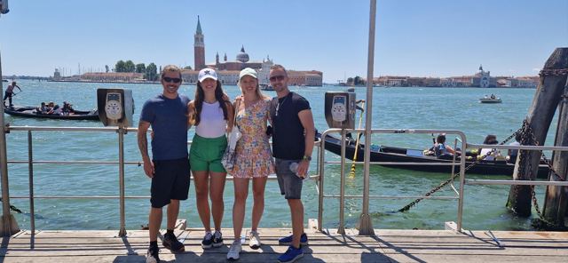 Four friends posing on a jetty with the lagoon and a bell tower behind.