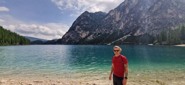 A man in a red T-shirt standing on the shore of a turquoise lake beneath mountains.
