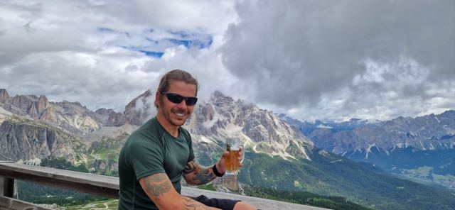 A man raising a beer at a mountain hut with dramatic Dolomite peaks behind.