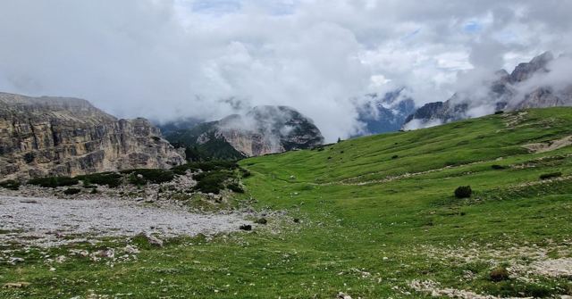 A green alpine meadow below rocky mountains with low clouds drifting across the peaks.