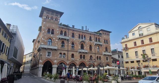 An ornate red-brick palazzo with arched windows overlooking a square with café tables.
