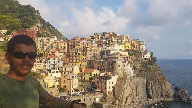 A man in sunglasses taking a selfie in front of the colourful clifftop houses of Manarola.