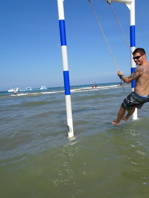 A man on a rope swing hanging between poles in the shallow sea.