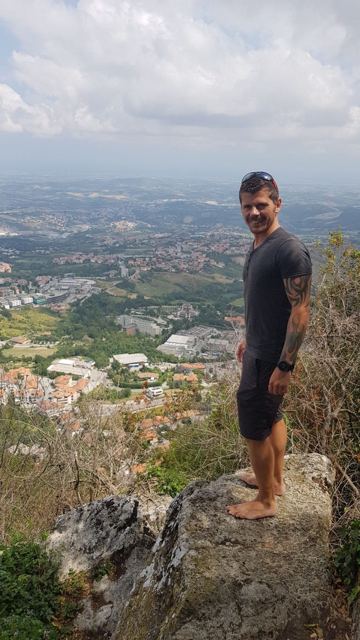 A barefoot man standing on a rocky outcrop above towns and hills in San Marino.