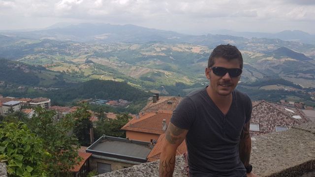 A man in sunglasses with rooftops and green hills stretching behind him.