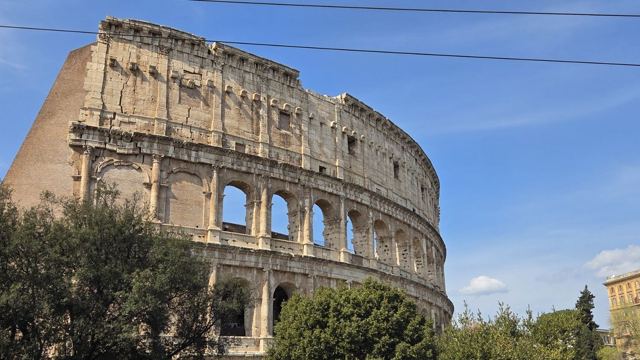 The curved stone arches of the Colosseum in Rome rising behind green trees under a blue sky.