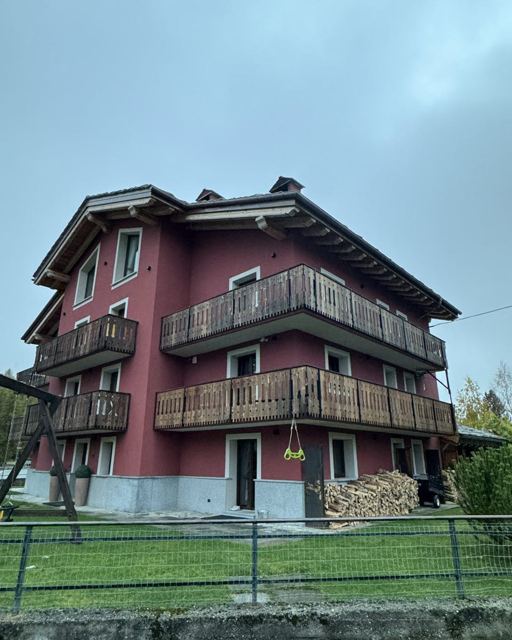 Red three-storey alpine house with wooden balconies and a green lawn under a cloudy sky.
