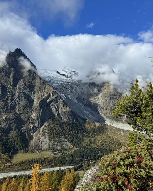 Jagged rocky peak beside glaciated mountains with autumn trees in the valley below.