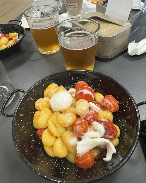 A pan of gnocchi with tomatoes and mozzarella beside two glasses of beer.