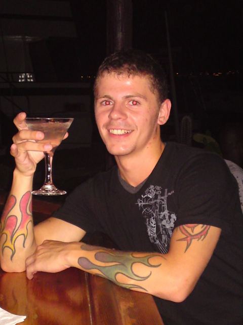 A smiling young man with tattooed arms holding up a martini glass at a bar.