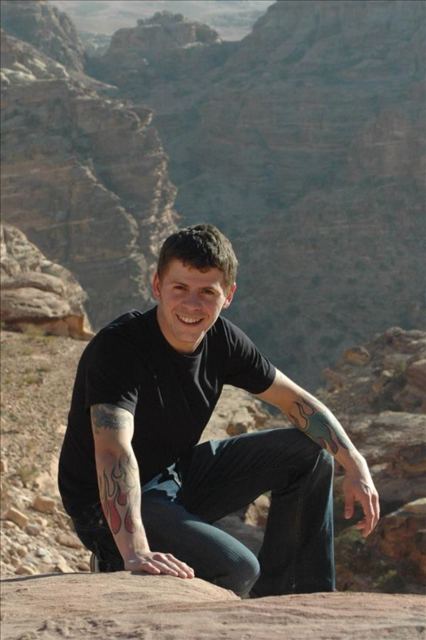 A smiling man in a black T-shirt crouching on sandstone rocks above a canyon at Petra.