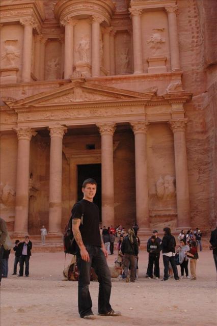 A man standing in front of the carved rose-red facade of the Treasury at Petra, with visitors around.