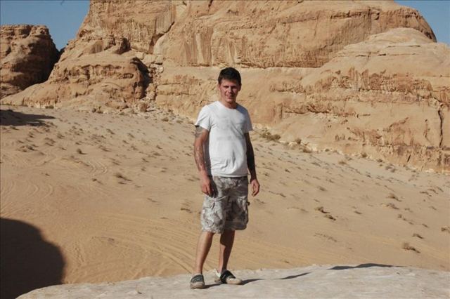 A man standing on red sand beside towering sandstone cliffs in the Wadi Rum desert.