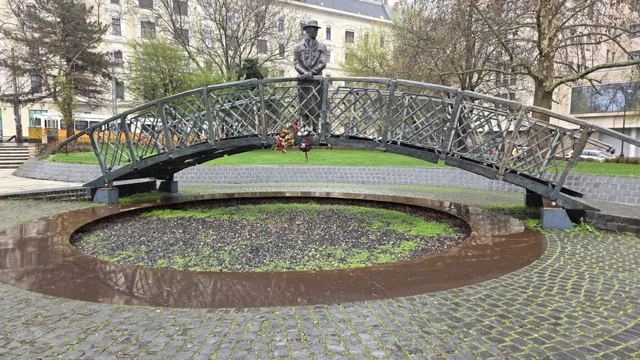 Bronze statue of a man standing on a small arched iron bridge over a round pool.
