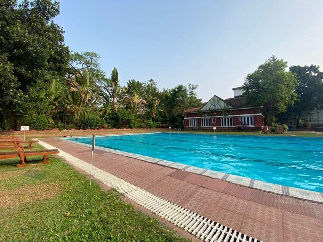 An outdoor swimming pool edged with brick paving, lawns, palm trees and a red-roofed building.