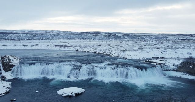 A wide waterfall flowing between snow-covered banks under grey sky.