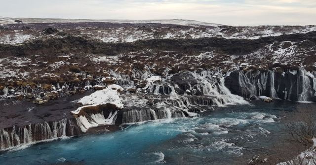 Many small waterfalls pouring from a dark lava field into a bright blue river.