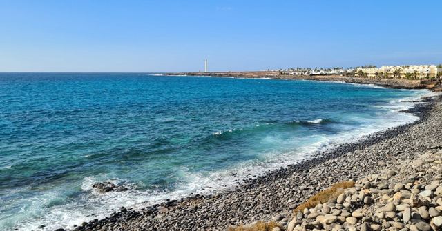 Deep blue sea breaking on a dark volcanic stone shore with a town in the distance.