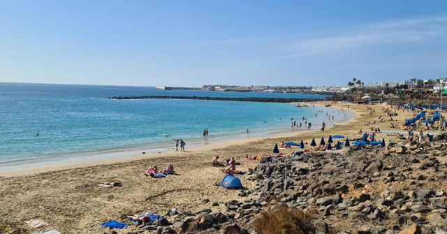 A sandy beach with turquoise water, sunbathers and dark rocks under a clear sky.
