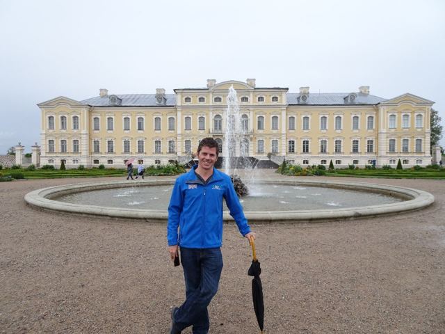 A man in a blue jacket holding an umbrella in front of a grand yellow baroque palace and fountain.