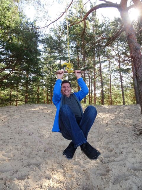 A man in a blue jacket on a rope swing among pine trees on sand.