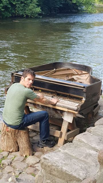 A man sitting at an old weathered grand piano on a riverbank.