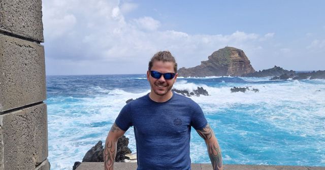 Smiling man in sunglasses and a blue T-shirt in front of crashing turquoise waves and a rocky islet.