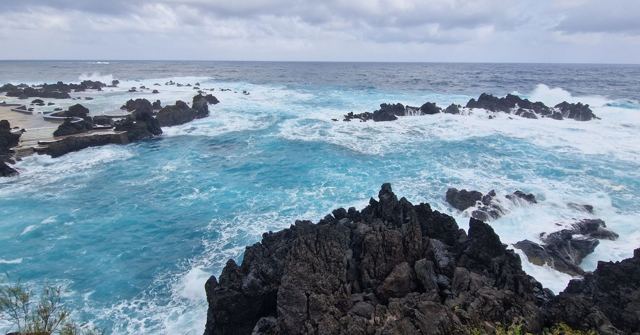 Turquoise waves crashing over black volcanic rocks along a rugged coastline.