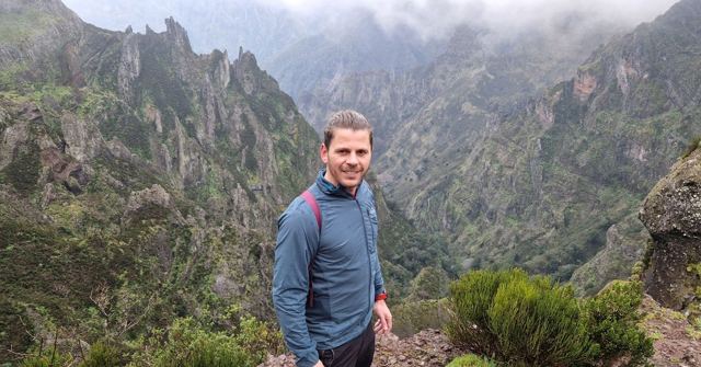 Man in a blue fleece standing in front of steep green mountain ridges and a misty valley.