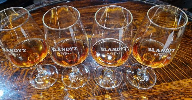 Four branded tasting glasses of Madeira wine in varying amber shades on a wooden table.