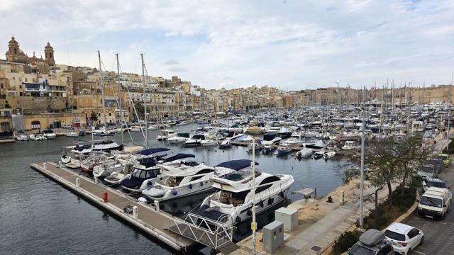 Marina full of white yachts beneath honey-coloured stone buildings and a church dome in Malta.