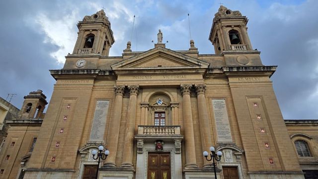 Front of a large golden-stone church with two bell towers and a columned facade under a blue sky.