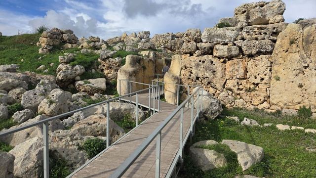 A boardwalk leading past huge ancient megalithic stone walls on a grassy hillside under blue sky.