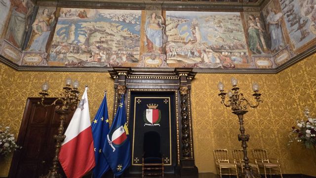 Ornate palace hall with painted frieze, yellow damask walls and Maltese and European Union flags.