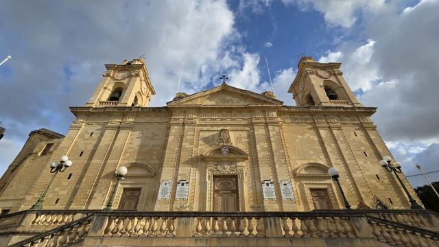 Wide-angle view of a golden limestone baroque church with two bell towers under a cloudy blue sky.