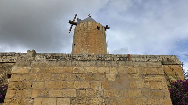 An old stone windmill tower rising above a golden limestone wall under a cloudy sky.