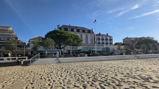 A wide sandy beach in Arcachon with seafront buildings and trees under a blue sky.