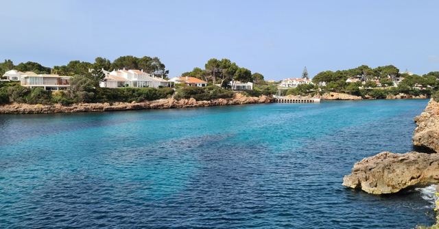 A turquoise cove with clear water and rocks, lined with white villas among pine trees.
