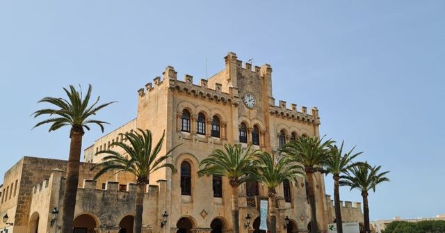 A golden stone building with battlements and a clock, fronted by palm trees under a blue sky.