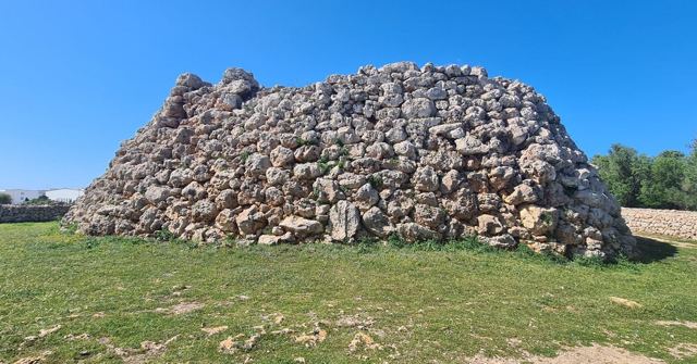 A large ancient stone mound built from rough rocks, standing on grass under a blue sky.