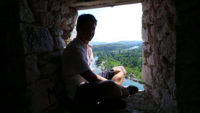 A man sitting in a stone window opening looking out at green countryside and a river.