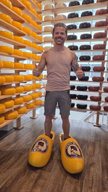 A man standing in giant yellow clogs in front of shelves of round Dutch cheeses.