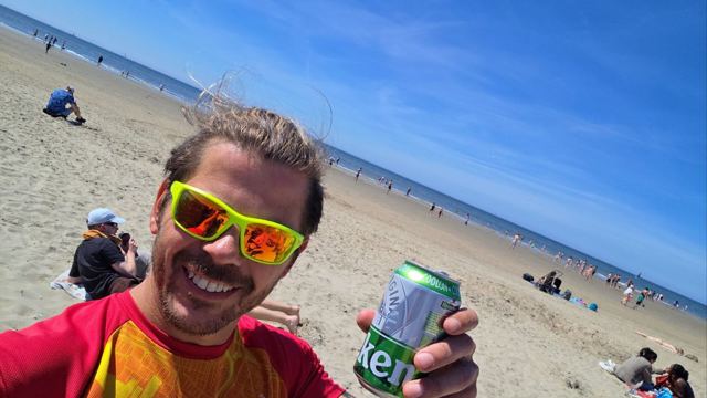 Smiling man in green sunglasses taking a beach selfie holding a green beer can, sea behind.