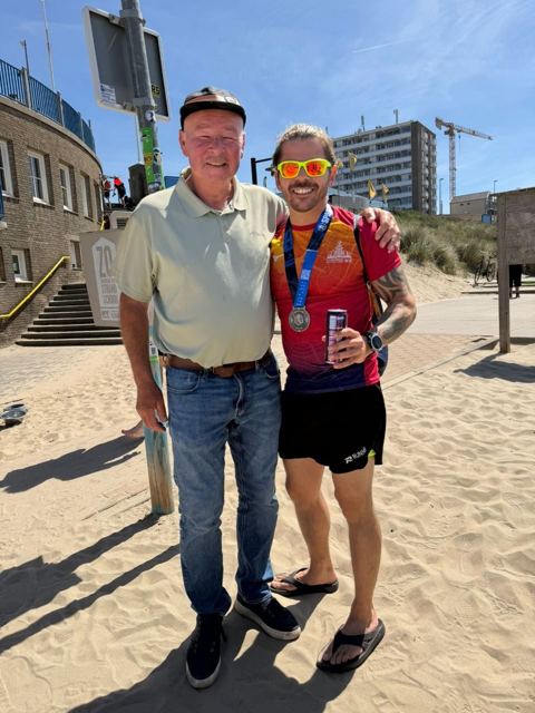 Two men standing on a sandy beach path, one wearing a race medal.