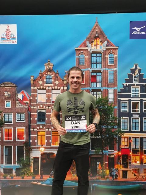 A man holding his race bib in front of a backdrop of colourful Amsterdam canal houses.