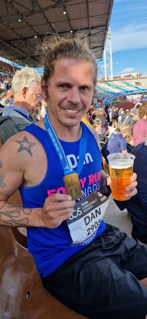 A smiling runner in a race vest holding a marathon medal and a cup of beer.