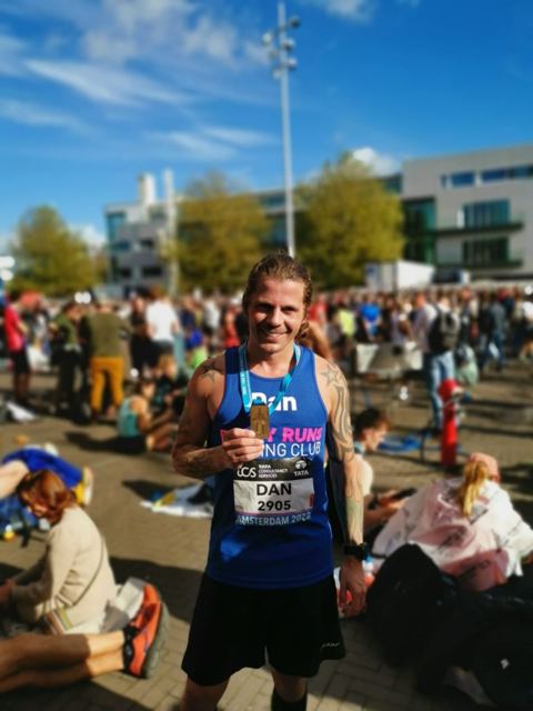 A runner with a race number and medal standing in a sunny crowd after the marathon.