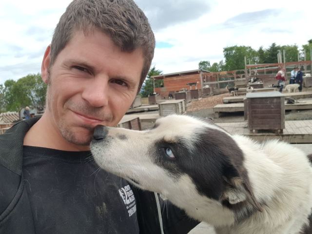 A man smiling as a black and white husky with pale blue eyes licks his cheek.