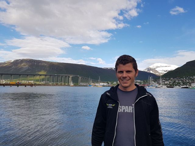A man standing by the water in Tromsø with the bridge and mountains behind.