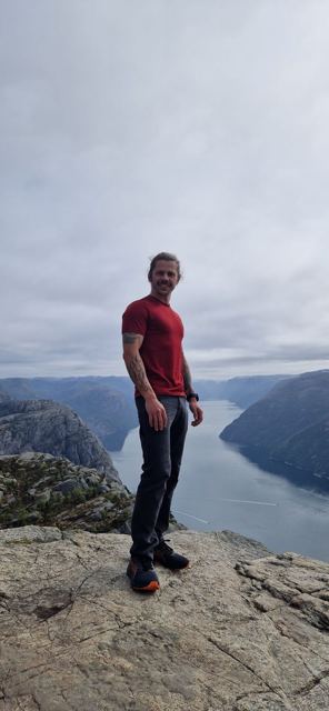 A man in a red T-shirt standing on a cliff edge above a long fjord.